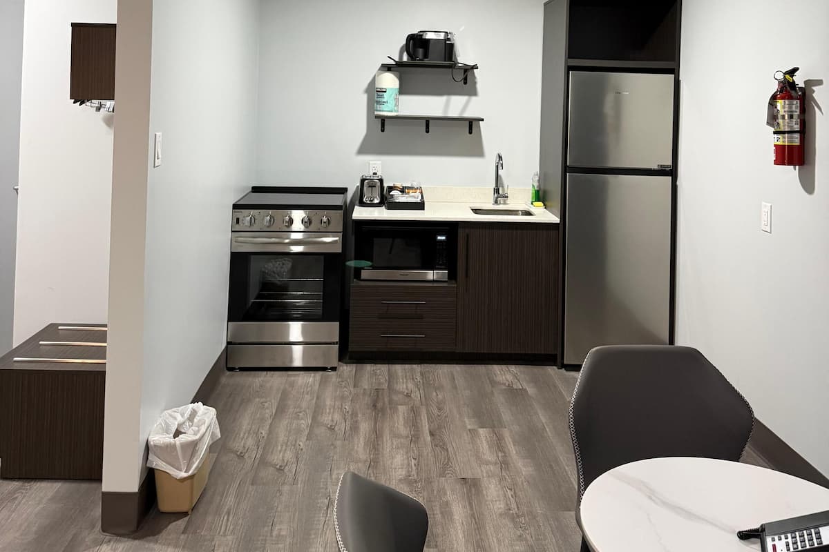 A compact kitchen with a stove, oven, microwave, and refrigerator. There is a dining table with chairs, a trash bin, and a fire extinguisher on the wall.