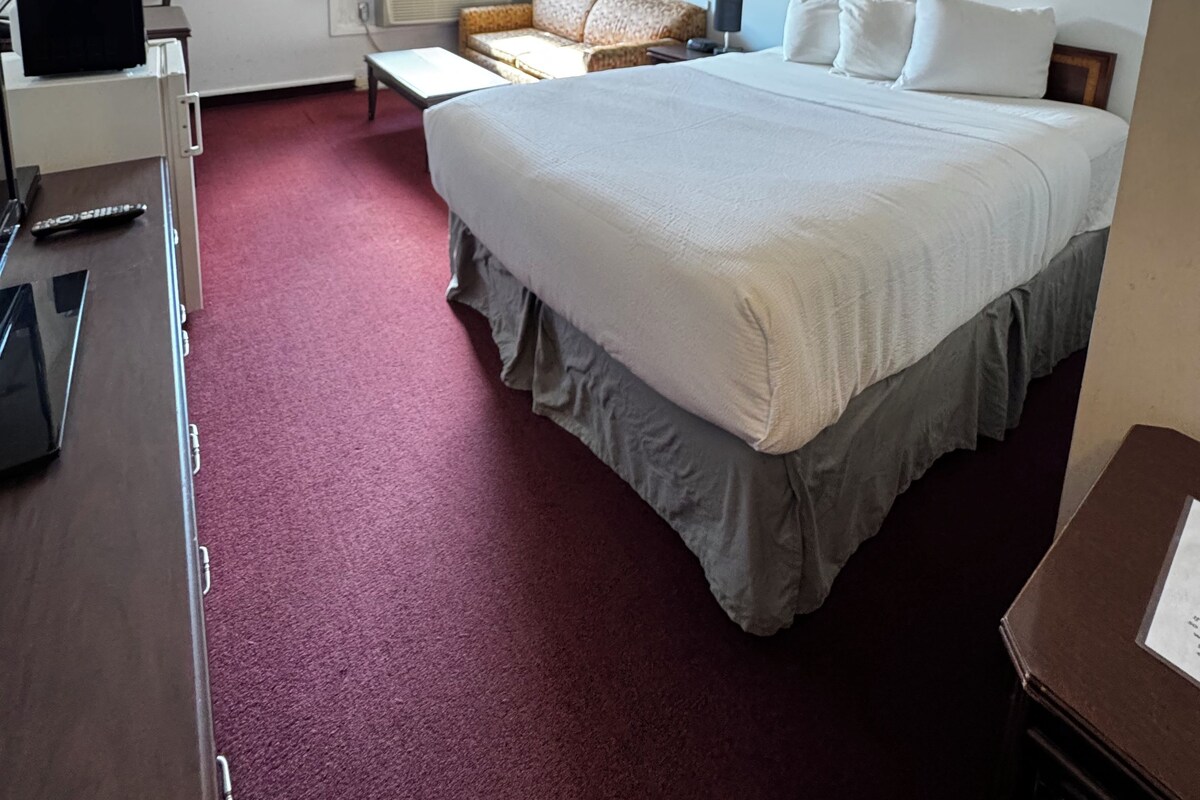 Habitació d'hotel amb un llit gran, una taula de treball amb una làmpada, una cadira i una finestra amb cortines.