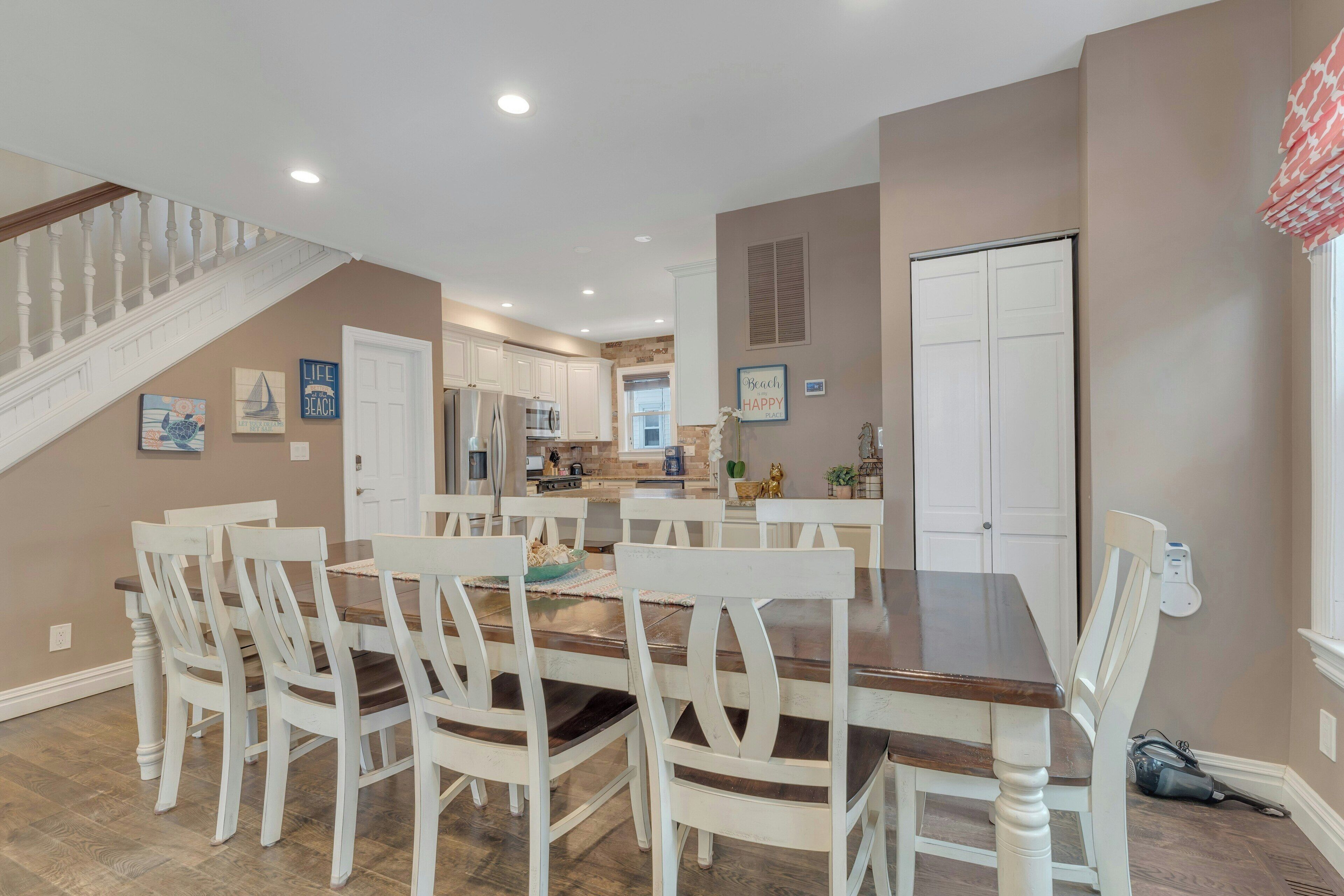 A dining area with a wooden table and white chairs, a kitchen in the background, and a staircase leading to another level.