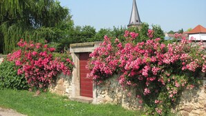 Un mur de pierre orné de plantes à fleurs roses et une porte rouge, qui donnent accès à un bâtiment doté d’un toit en forme de flèche.
