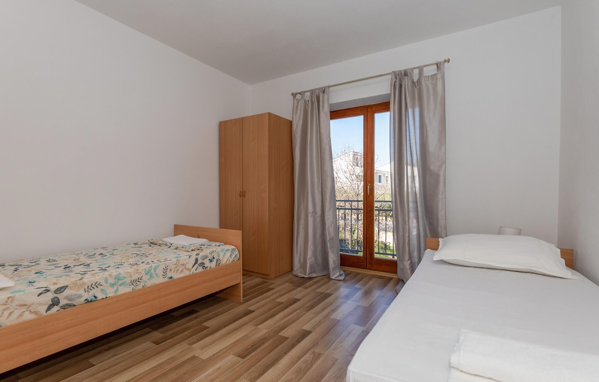 A bedroom with two beds, wooden flooring, a wardrobe, and a balcony with metal railings.