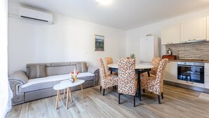 A modern living room with a grey sofa, a small white table, and patterned chairs. A kitchen with white cabinets and a stove is visible in the background. - 3 bedroom cozy home in Kremena (Kremena)