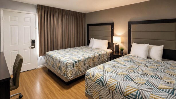 Habitación de hotel con dos camas, un escritorio, una silla, una puerta y una mesita de noche con lámpara.