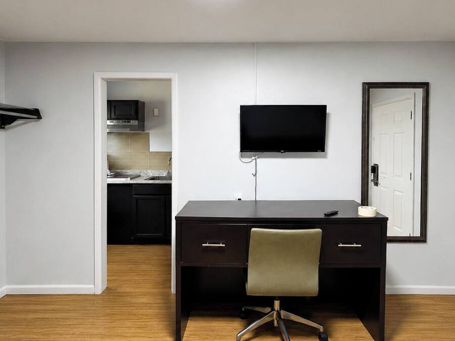 A room with a desk, chair, TV, and a door leading to a kitchen.