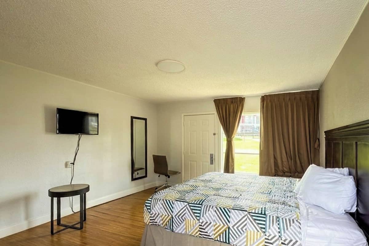 Quarto de hotel com uma cama, televisão na parede, mesa redonda pequena, uma cadeira e uma porta.