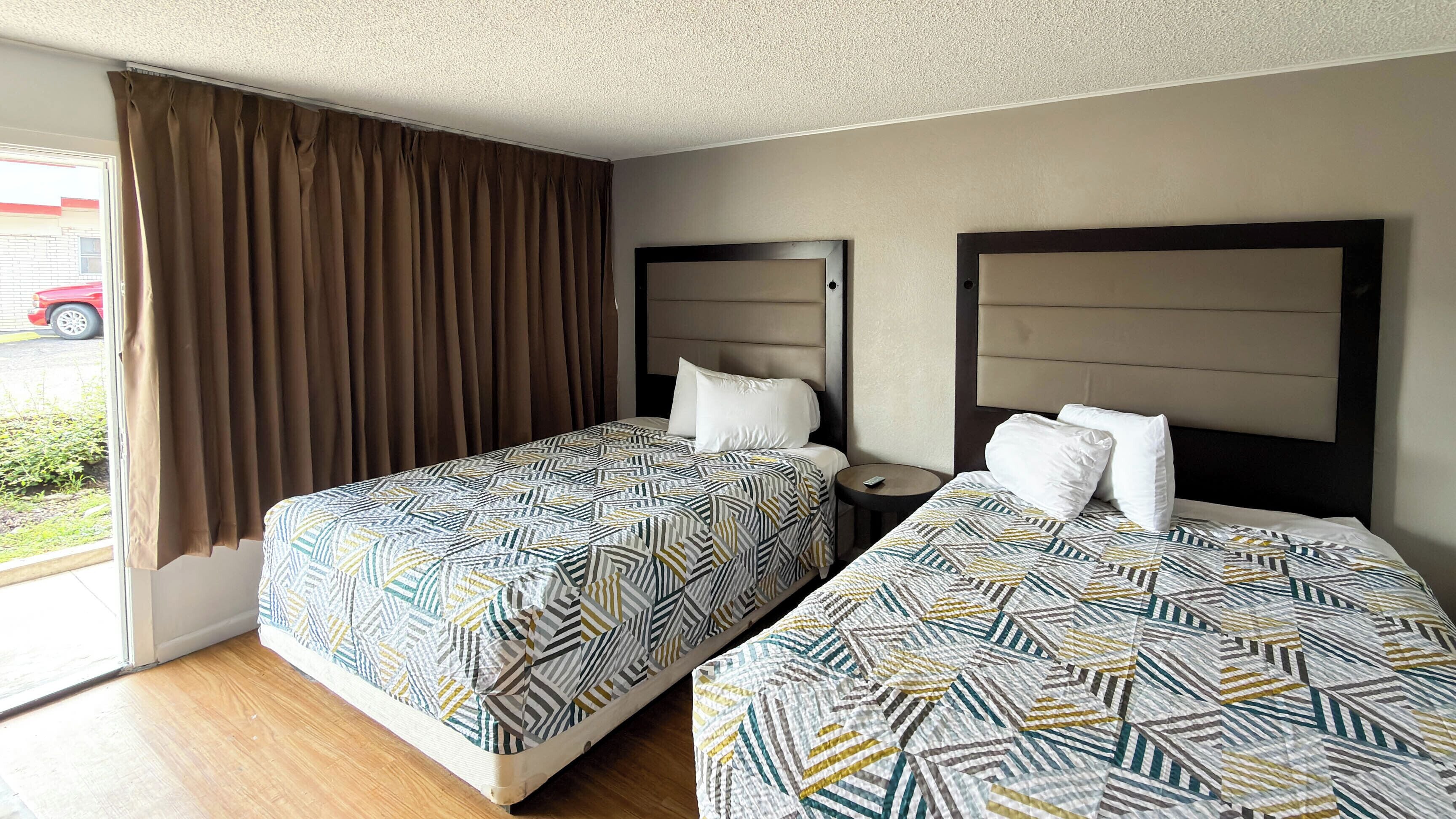A hotel room with two beds, a window with curtains, a bedside table, and a view of the street outside.