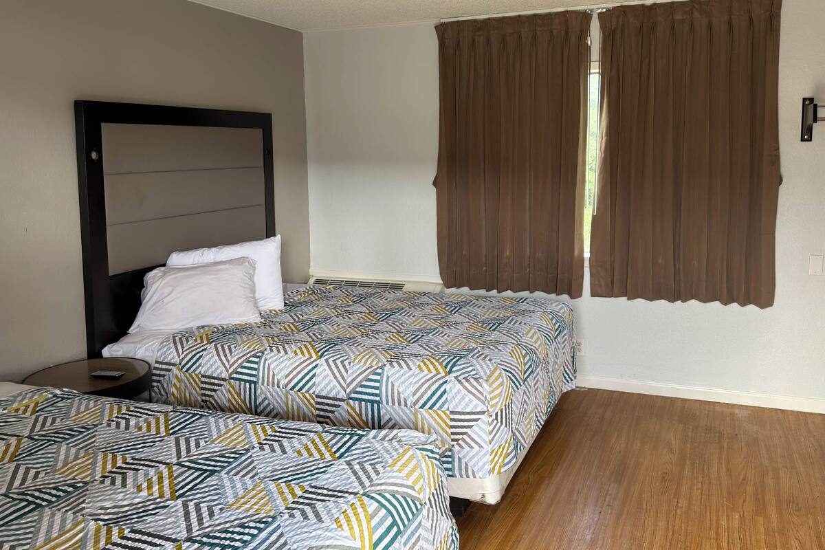 Quarto de hotel com duas camas, uma mesa redonda pequena, cabeceira e janela com cortinas castanhas.