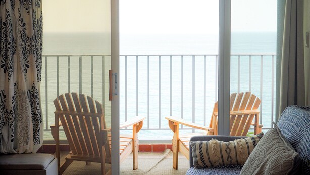 A balcony with wooden chairs and a sofa, overlooking a body of water. - Modern 1-Bedroom in the Heart of Condado with Stunning Ocean Views (San Juan)