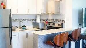 A kitchen with white cabinets, a microwave, a stove, and two wooden chairs. - Modern 1-Bedroom in the Heart of Condado with Stunning Ocean Views (San Juan)