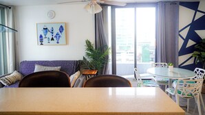 A modern living room with a ceiling fan, a sofa, a dining table, and chairs. There is a large window with curtains, a plant, and a wall with geometric patterns. - Modern 1-Bedroom in the Heart of Condado with Stunning Ocean Views (San Juan)