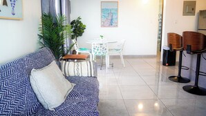 A modern living room with a sofa, a dining table with chairs, and a kitchen area with bar stools. - Modern 1-Bedroom in the Heart of Condado with Stunning Ocean Views (San Juan)