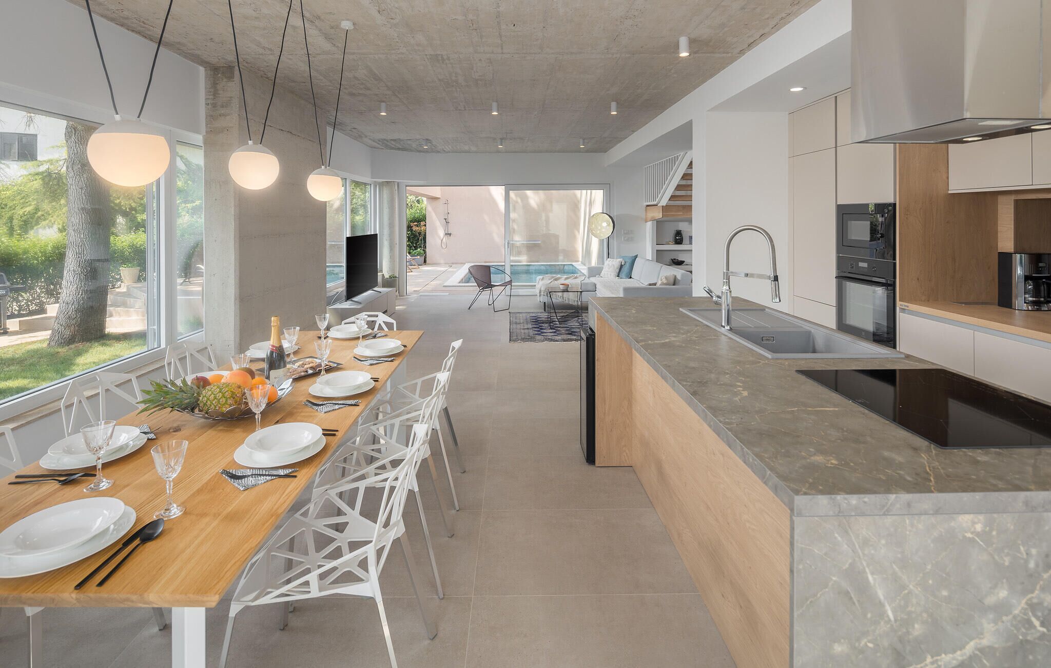 A modern kitchen with a central island, a dining area with a long table, and a view of a pool and outdoor seating.