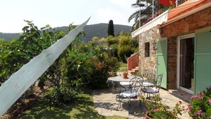 Um edifício de pedra com uma persiana verde, um pátio com mesa e cadeiras, e um jardim com diversas plantas e uma vela branca grande.
