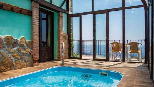 A swimming pool with a view of the sea, a glass-enclosed patio with chairs, and a stone wall.