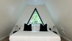 Una stanza triangolare con un letto al centro, sotto una grande finestra, biancheria da letto bianca, cuscini neri e comodini.