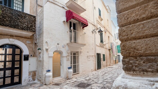 Una stretta strada acciottolata fiancheggiata da edifici con balconi, una porta bianca e un tendone rosso.