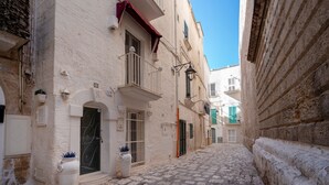 Una stretta via acciottolata fiancheggiata da edifici storici con balconi, un cielo azzurro limpido e una porta bianca con una targa numerata.