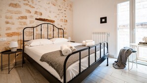 A bedroom with a stone wall, a bed with white linens, a bedside table with a lamp, and a view through a window.