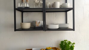 A kitchen with a black metal shelf unit holding glassware and dishes, a wooden tray with food items, and a potted plant.