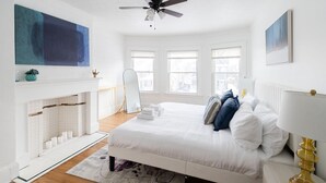 Une chambre avec un lit blanc, un ventilateur de plafond, une cheminée et de grandes fenêtres.
