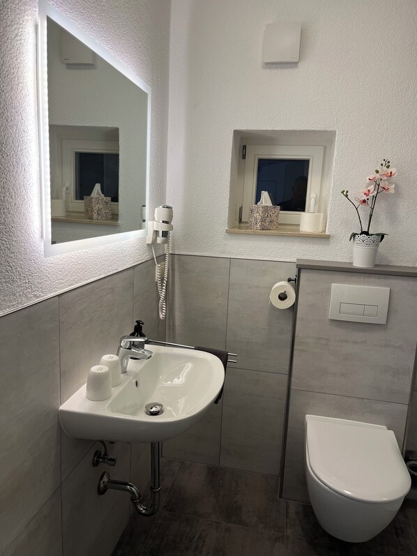 Un baño moderno con un lavabo blanco, un inodoro, un espejo y una ventana pequeña con una planta.