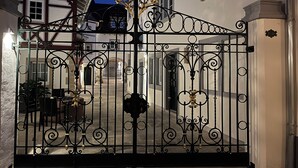 Una reja negra de hierro forjado con diseños intrincados, que da acceso a un edificio con paredes blancas y numerosas ventanas.