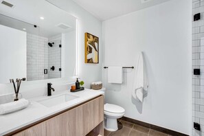 A modern bathroom with a white sink, black faucet, and a large mirror. There is a toilet with a towel rack and a painting on the wall.