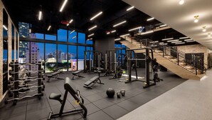 A modern gym with various exercise equipment, large windows offering city views, and a staircase leading to an upper level.