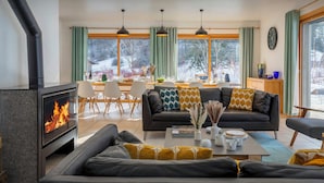 A modern living room with a wood-burning stove, a sofa, a dining area, and large windows with a view of trees.