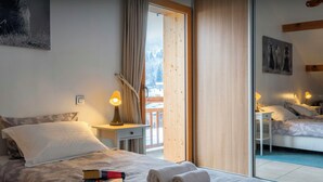 A hotel room with a bed, a bedside table with a lamp, a small desk, and a large window with a view of trees and mountains.