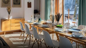 A dining area with a wooden table set for six, white chairs, and a view of the outdoors through large windows.
