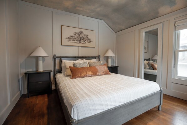 A bedroom with a large bed, two nightstands, a framed picture on the wall, and a window with a view.