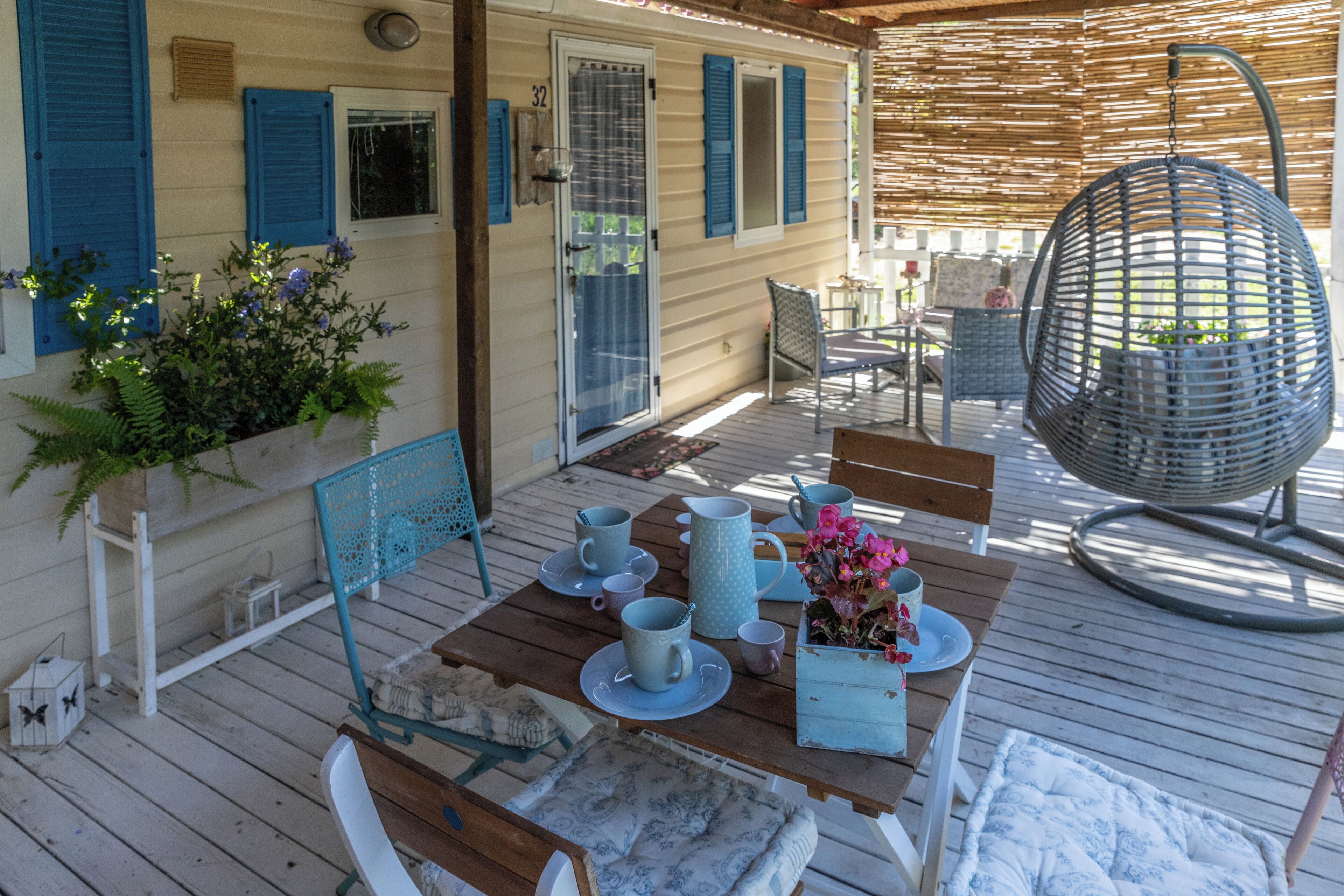 Um deck de madeira com mesa posta para refeições, cadeiras, uma cadeira suspensa e um vaso com planta.
