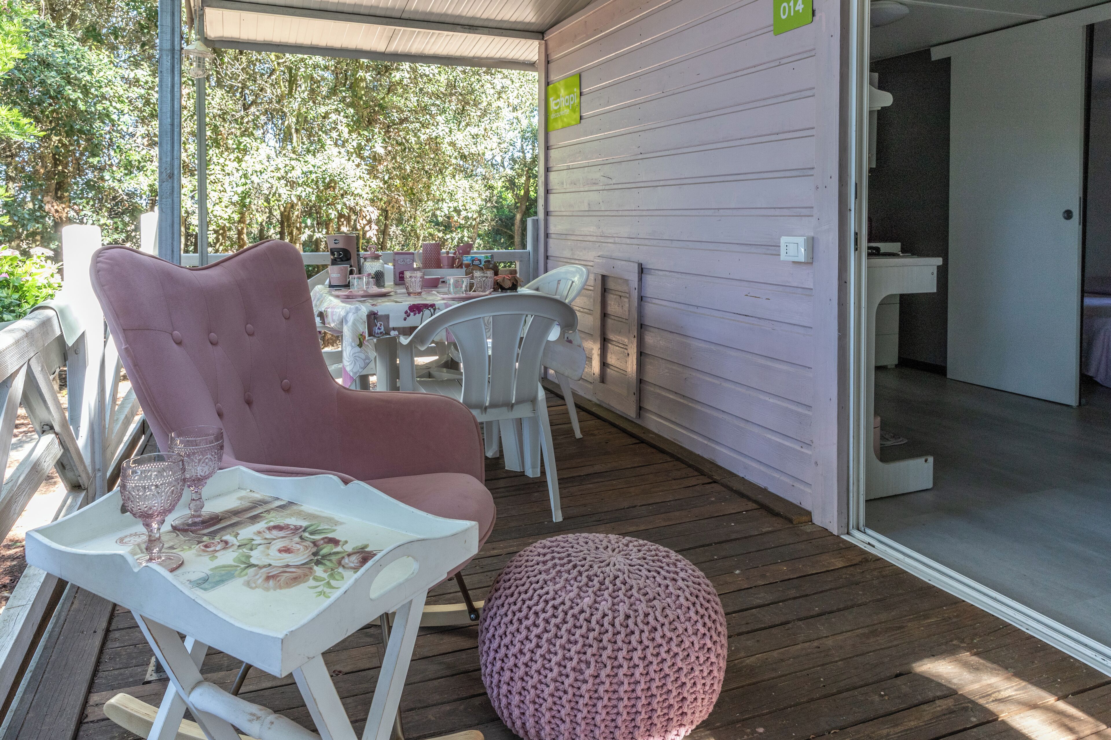 Un espace extérieur abrité, avec une table et des chaises, un pouf rose et un panneau indiquant le numéro de chambre : 014.