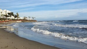 A beach with waves, a sandy shore, and a building with white walls and palm trees in the background.
