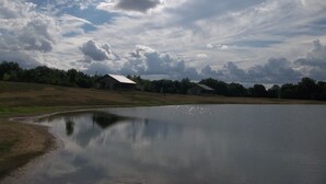 A calm lake with a reflection of the sky and clouds, surrounded by grassy areas and two small buildings with sloped roofs.