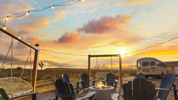 A fire pit surrounded by Adirondack chairs, a swing set, and a bus in a desert landscape during sunset. - Joshua Tree-Spa Pool Firepit Sauna Game Room (Yucca Valley)