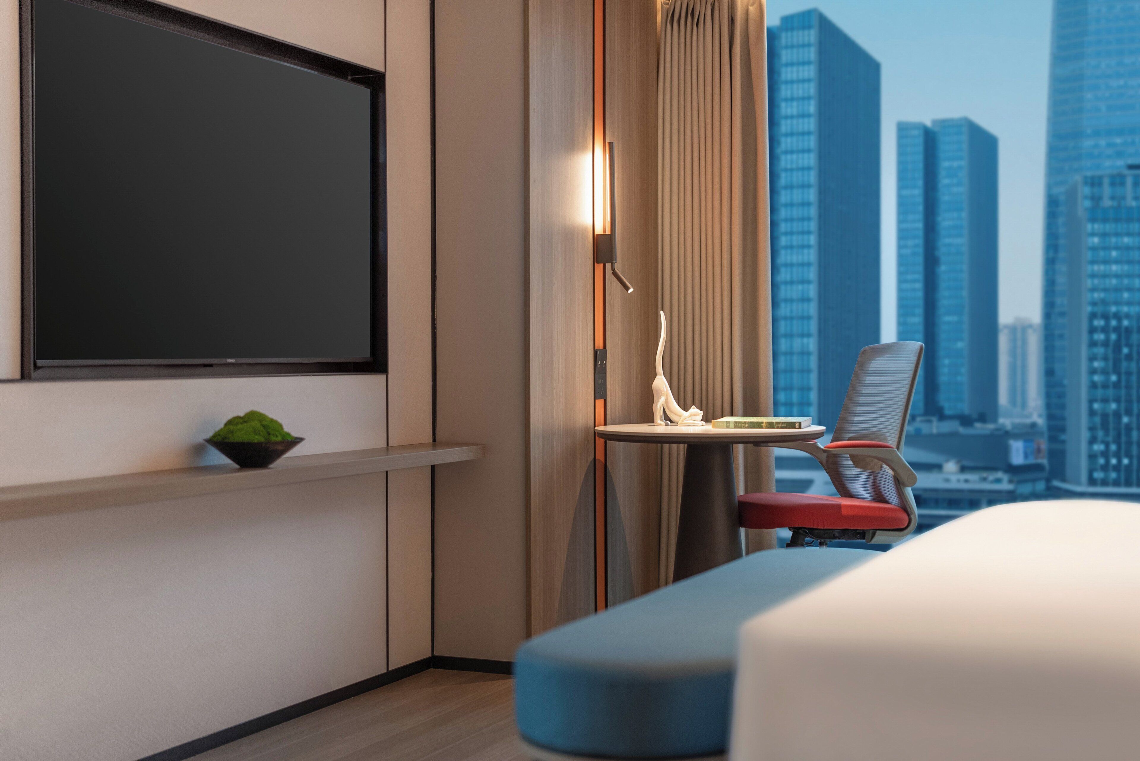 Ett modernt hotellrum med en stor platt-TV, ett litet bord med en dekorativ detalj, en stol och utsikt över en stadspanorama.