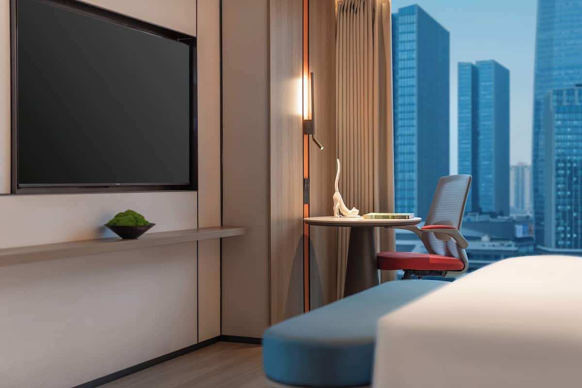 A modern hotel room with a large flat-screen TV, a small table with a decorative item, a chair, and a view of a cityscape.