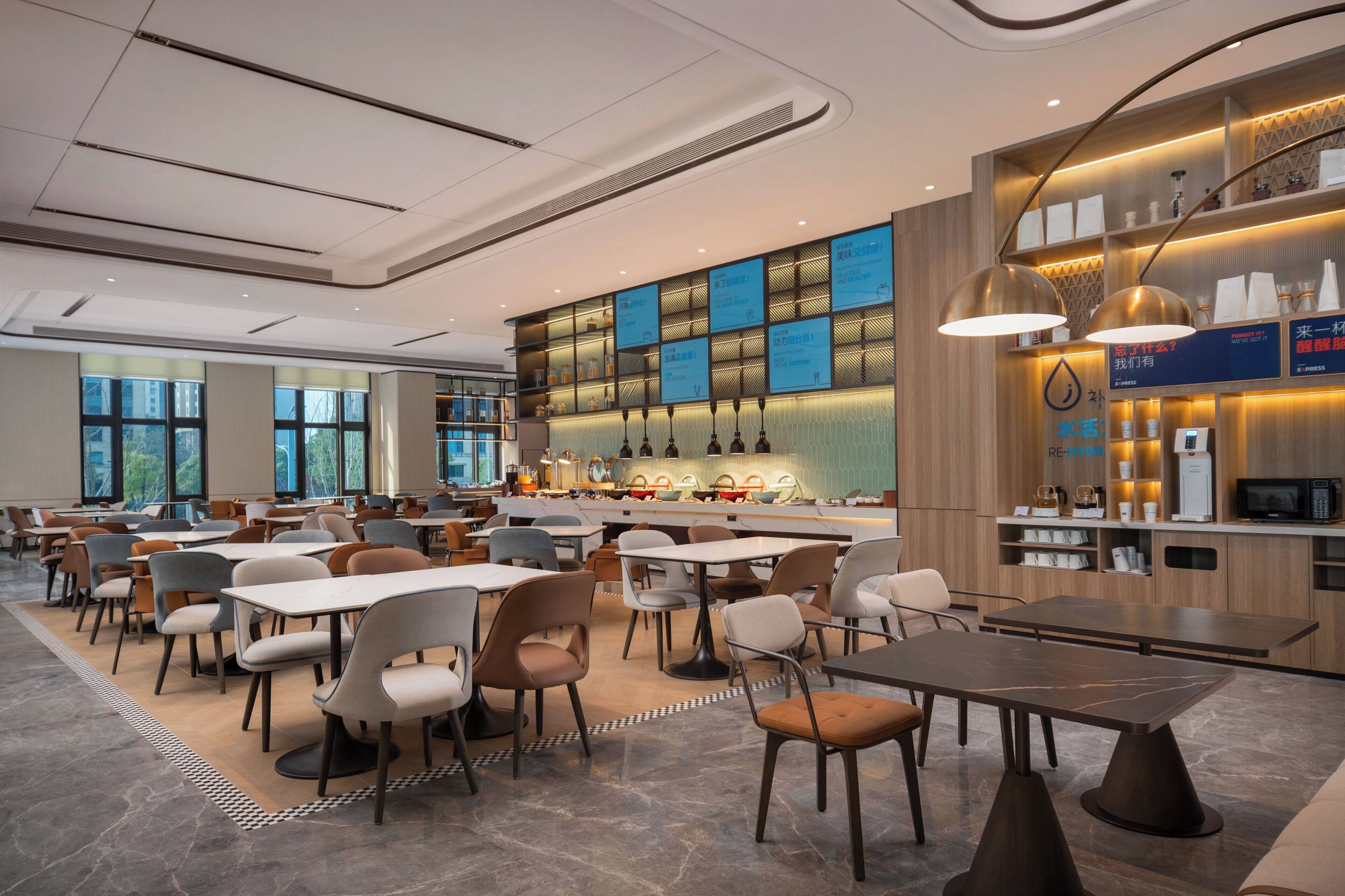Un comedor moderno con varias mesas y sillas, una zona de bar con estanterías e iluminación, y amplias ventanas que ofrecen luz natural.
