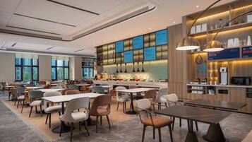 Un comedor moderno con varias mesas y sillas, una zona de bar con estanterías e iluminación, y amplias ventanas que ofrecen luz natural.