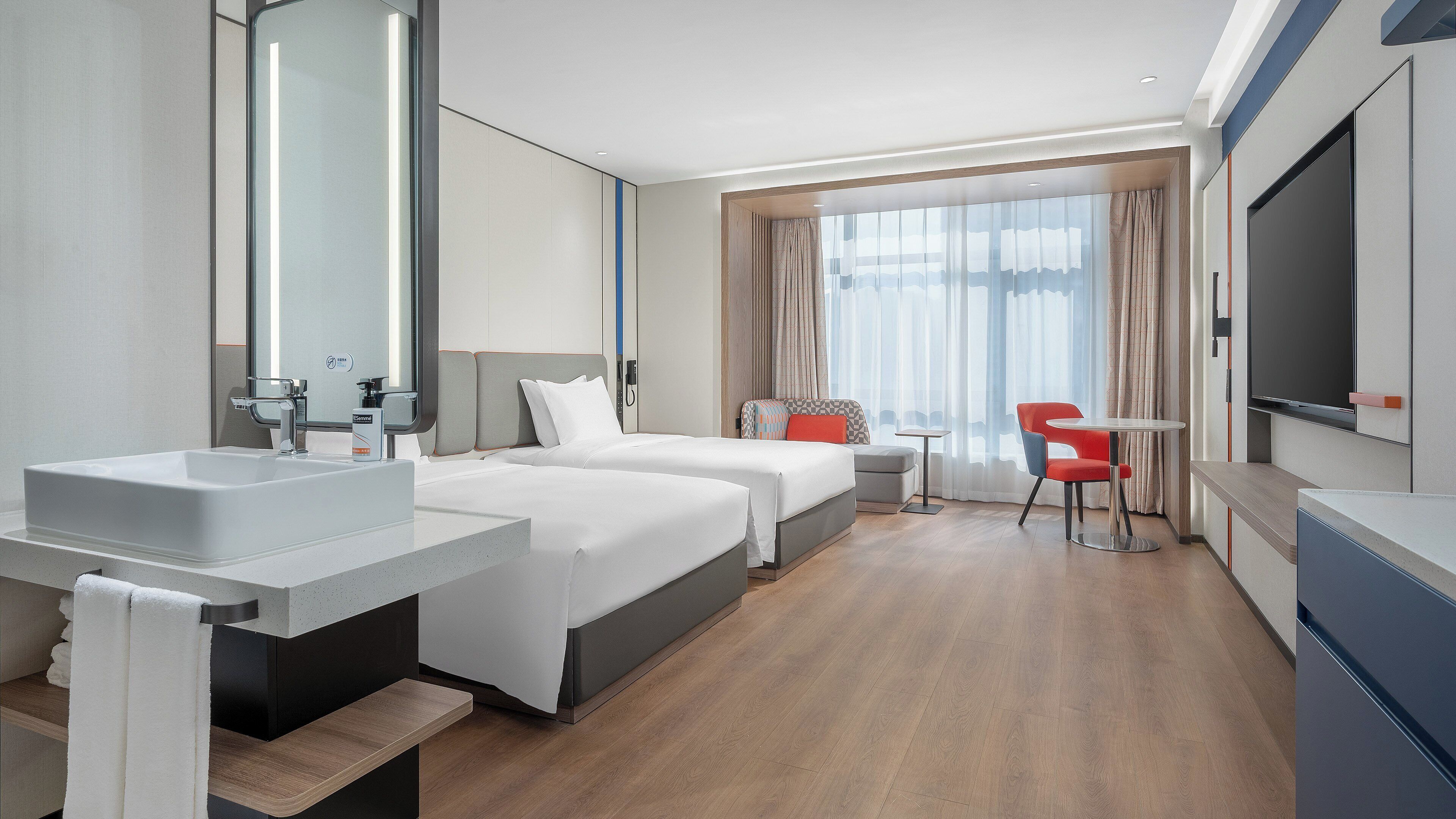 A modern hotel room with two beds, a bathroom with a sink, a small table, a chair, and a TV.