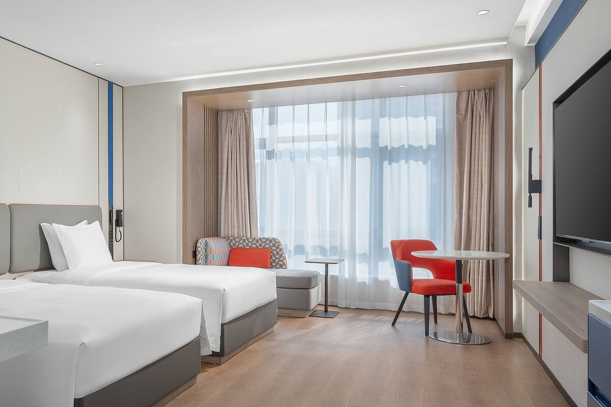 A hotel room with two beds, a small table, a chair, a TV, and a large window with curtains.