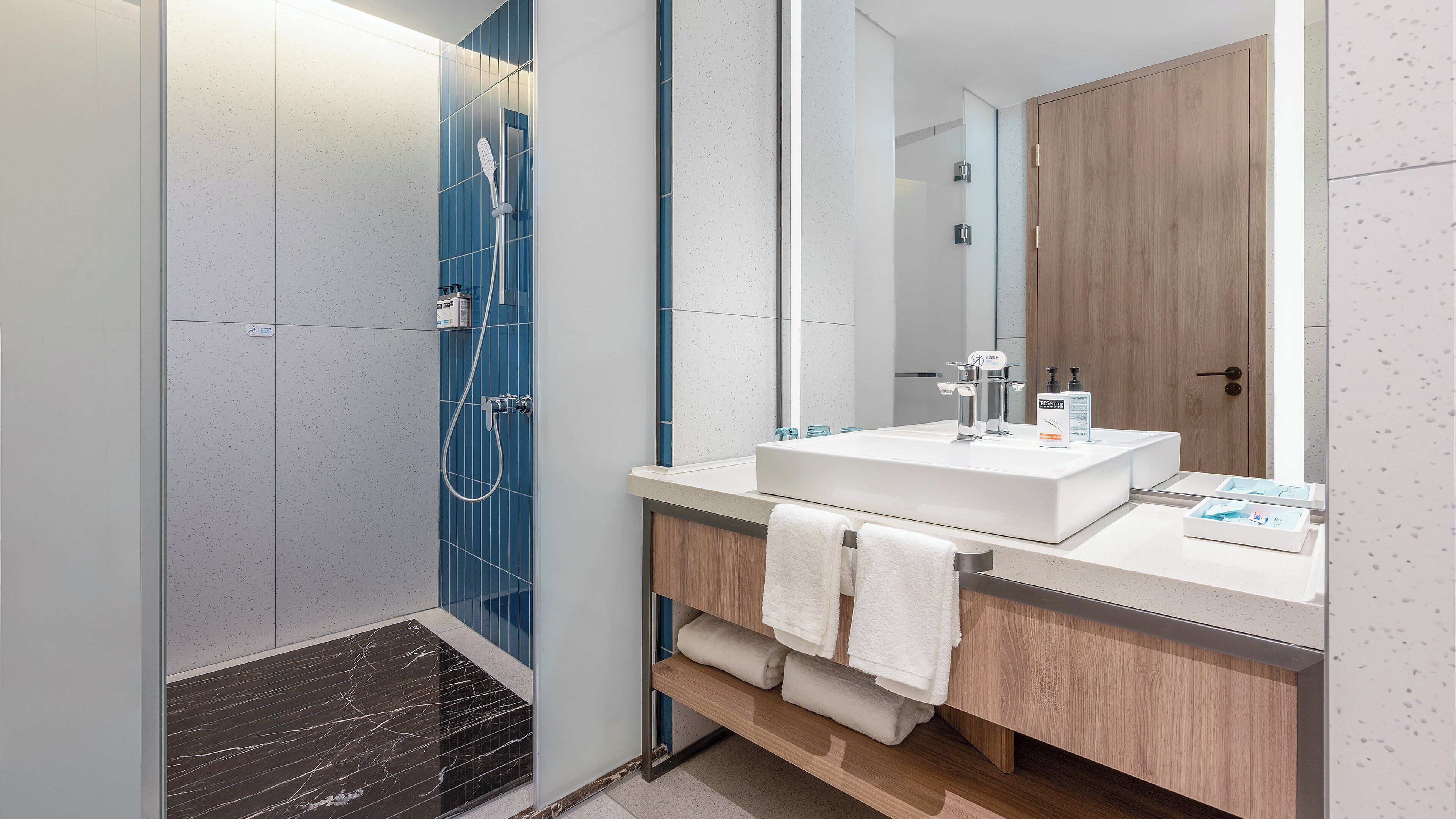 A modern bathroom with a large mirror, two sinks, a shower, and a wooden cabinet.