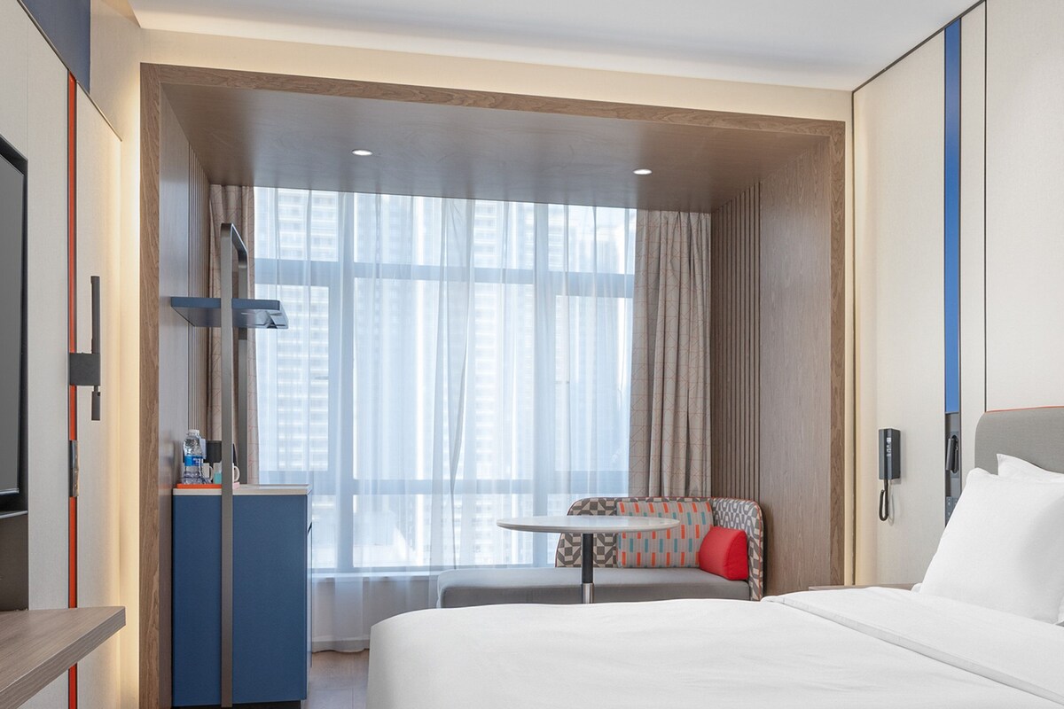 Ein Hotelzimmer mit einem großen Bett, einem Schreibtisch, einem Sessel und einem fenster mit Vorhängen.