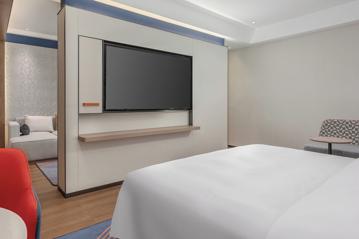 Ein modernes Hotelzimmer mit einem großen Bett, einem an der Wand montierten Fernseher, einer Sitzecke mit einer gemusterten Bank und einem kleinen Tisch.