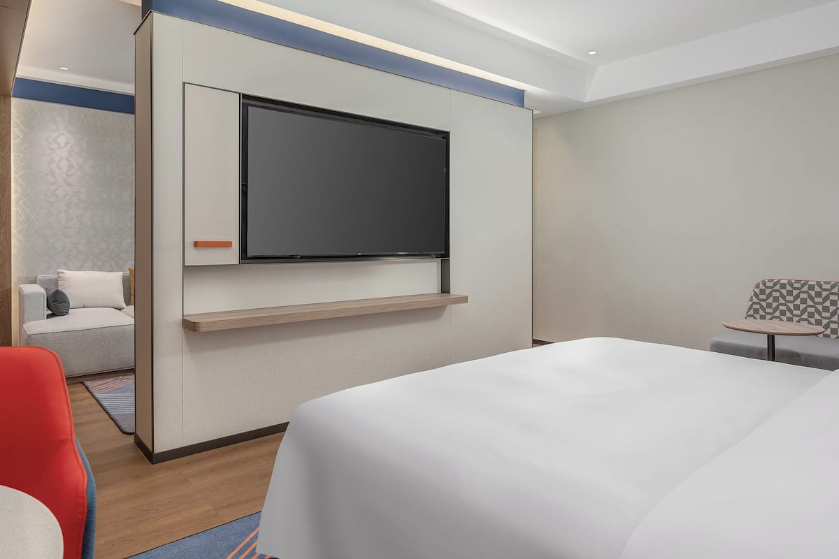 A modern hotel room with a large bed, a TV mounted on the wall, a seating area with a patterned bench, and a small table.