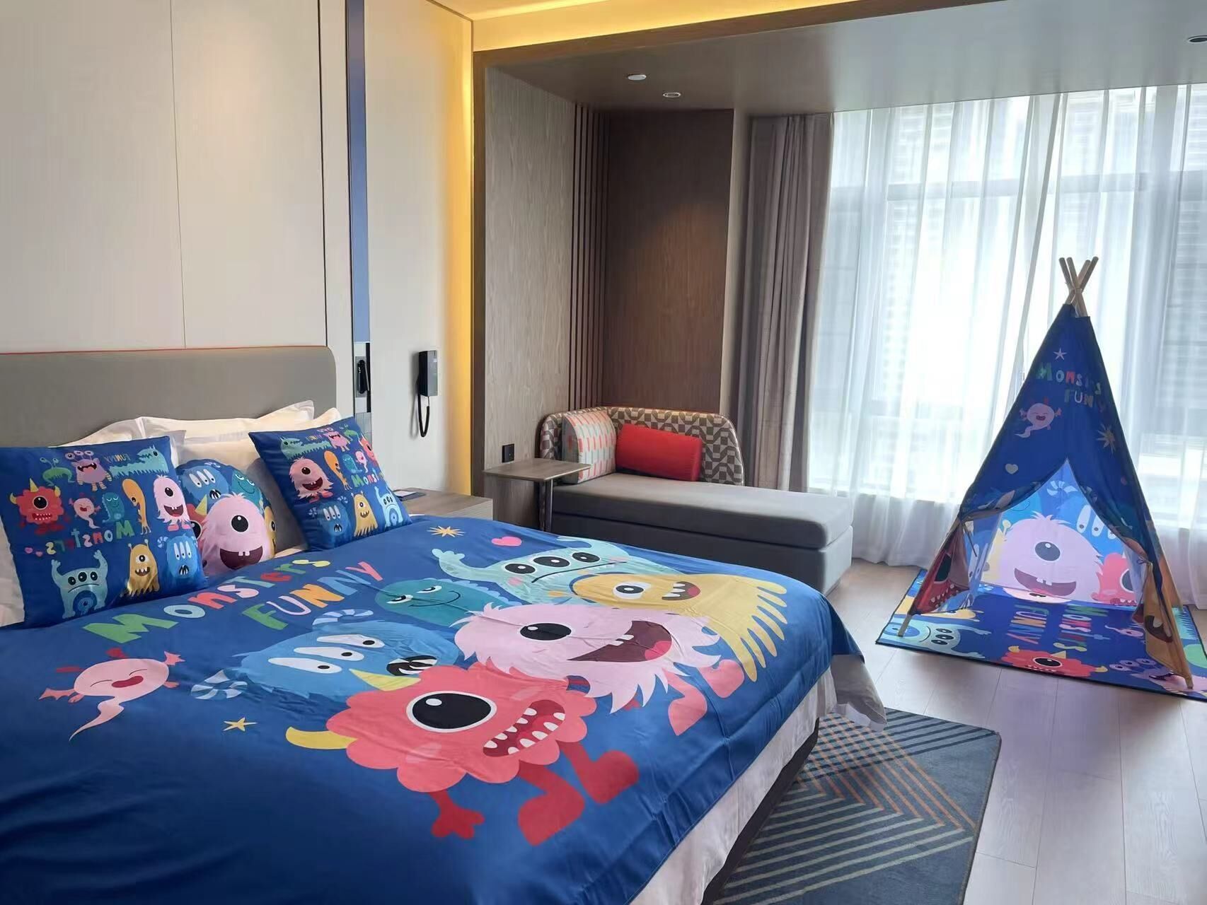 A hotel room with a bed featuring a blue bedspread with cartoon characters, a small sofa, a window with curtains, and a tent-like decoration with cartoon characters.