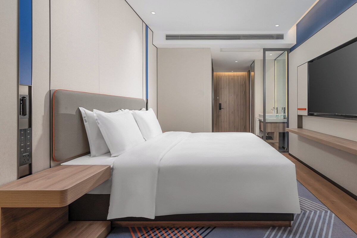 Ein modernes Hotelzimmer mit einem großen Bett, einem Nachttisch, einem integrierten Fernseher und einem durch eine offene Tür einsehbaren Badezimmer.