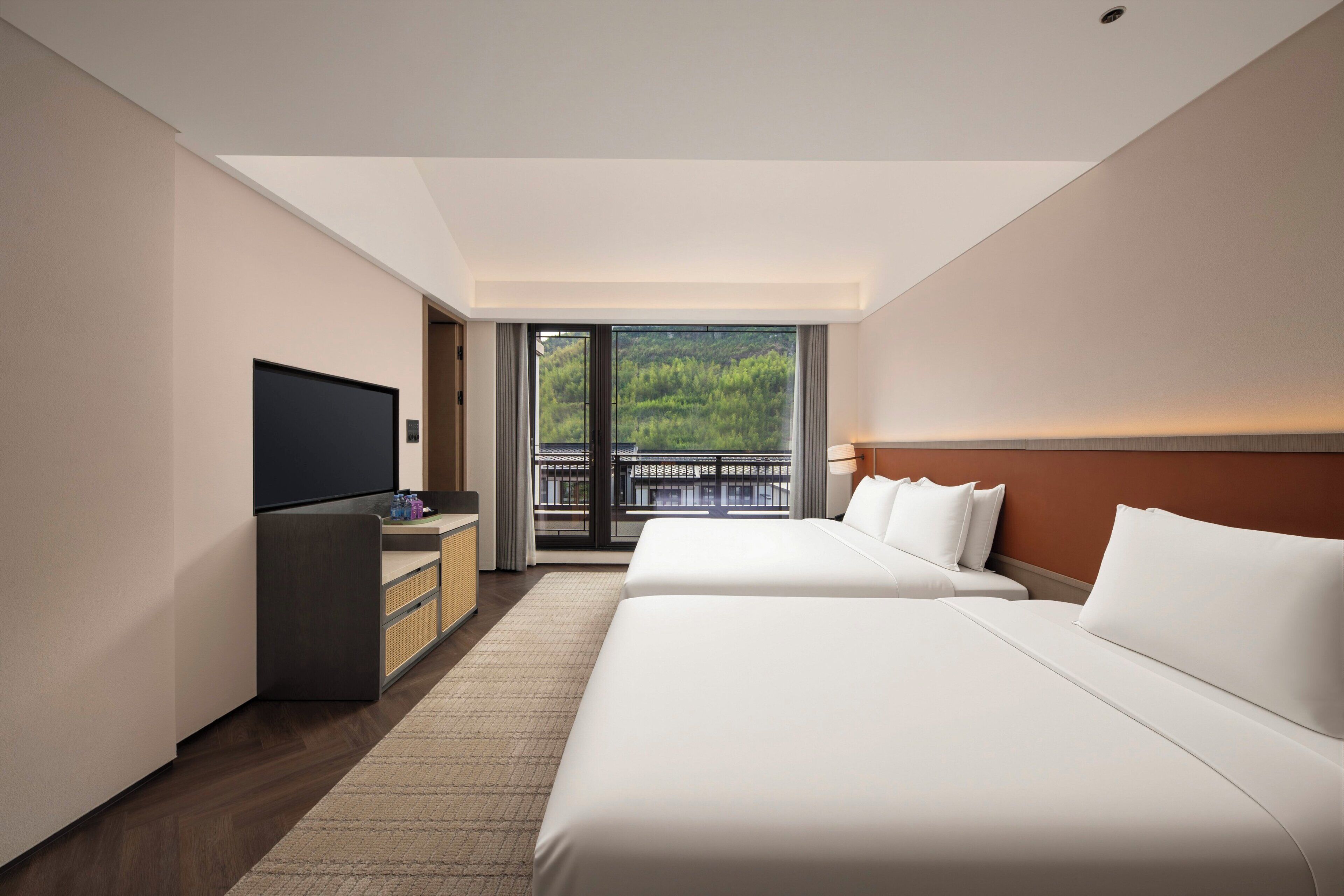 A hotel room with two beds, a TV, a desk, and a balcony with a view of greenery.